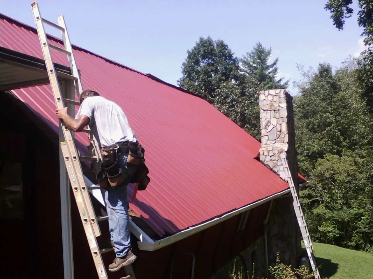 Roofer on a ladder beside a red metal roof and stone chimney — Soffit & Fascia Repair by Roofing Brothers in Tuskegee